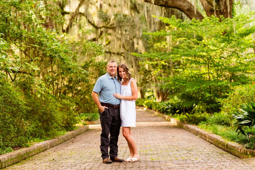 Couple posing on brick path surrounded by lush green trees draped with Spanish moss in a scenic garden.