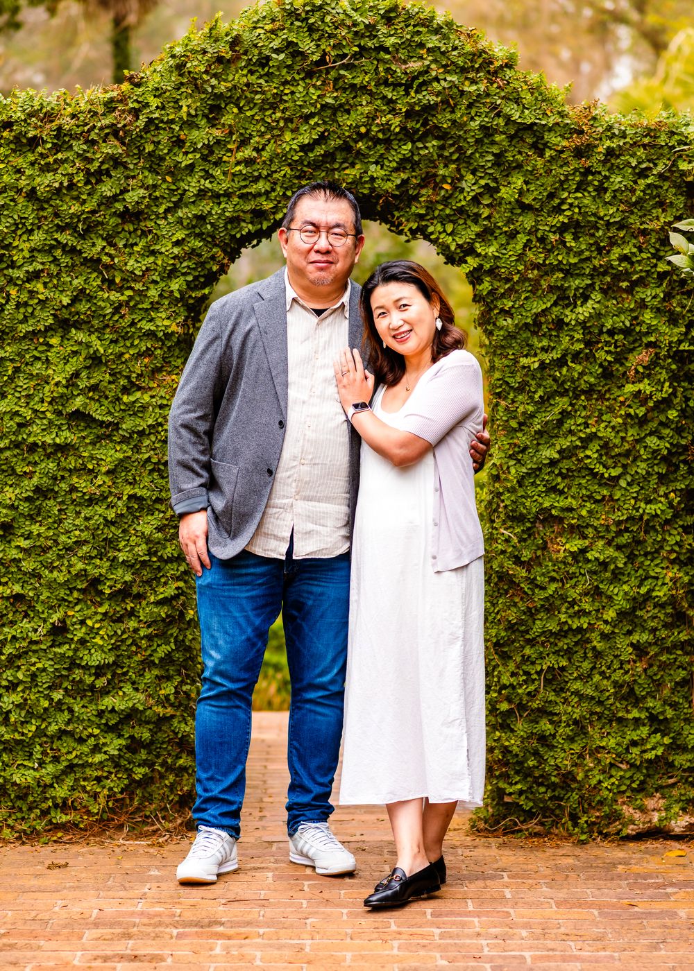 A couple poses together against a lush green hedge backdrop on a brick pathway in an outdoor garden setting.