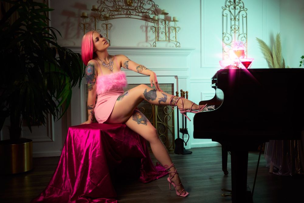 Editorial portrait of a tattooed pink-haired woman in a pink dress and heels seated beside a black grand piano with a glowing cocktail prop.