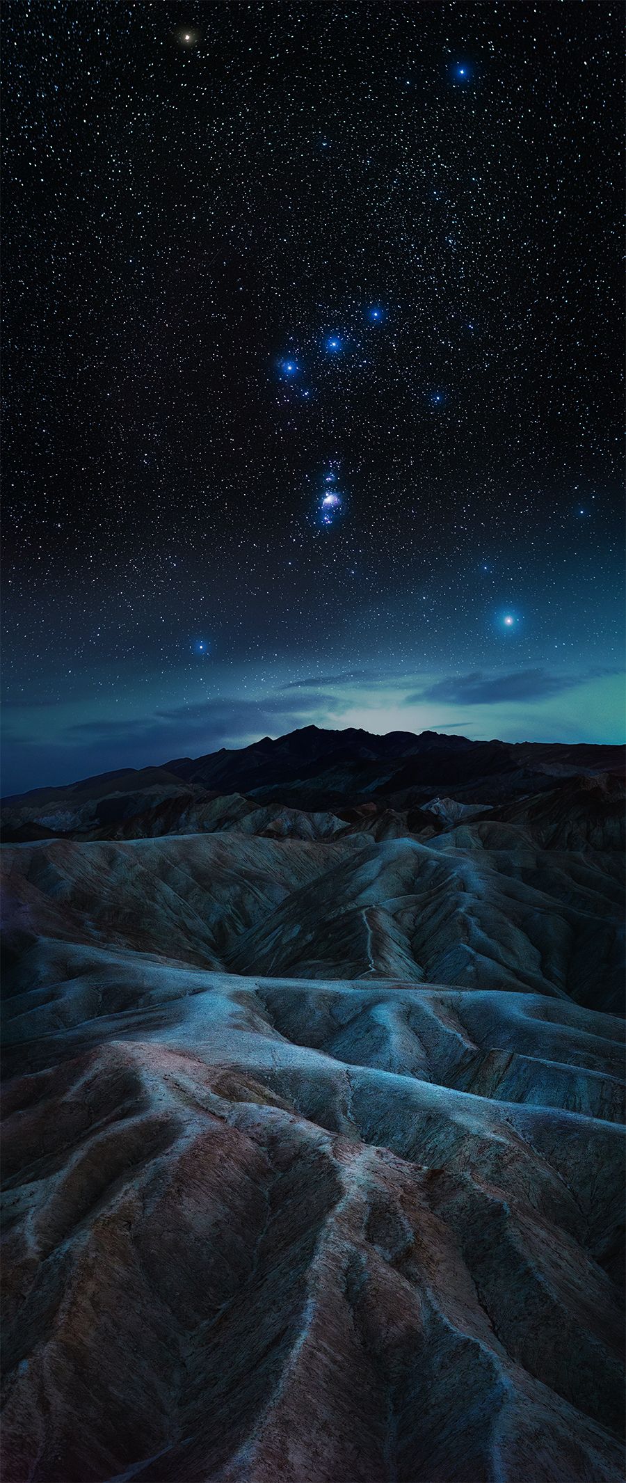 Dramatic night landscape of Death Valley's rugged badlands under a starry blue sky with mountains silhouetted against the horizon.