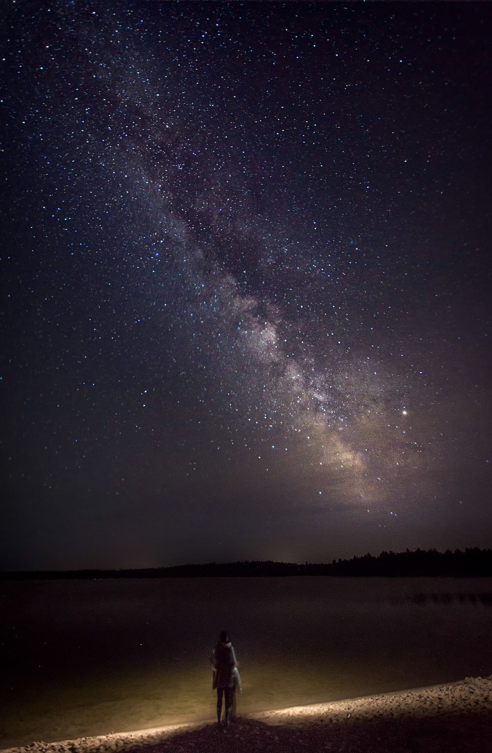 Silhouette stands beneath brilliant display of Milky Way stars over dark landscape.