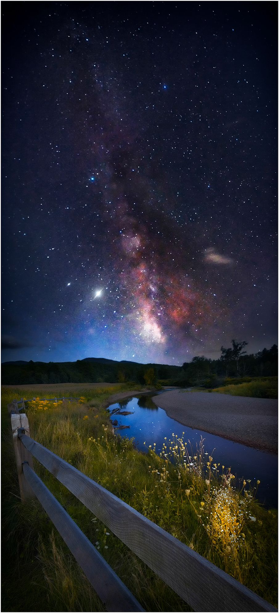 Milky Way galaxy stretches across night sky above a wooden fence and reflective stream.