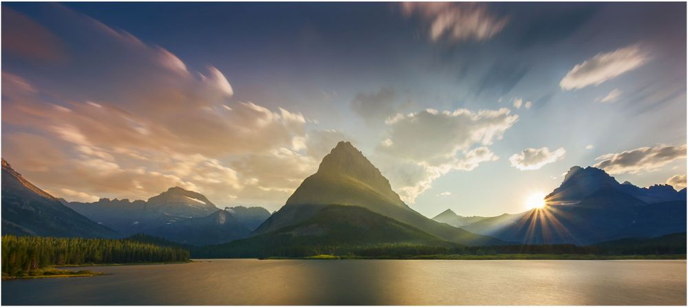 Mountain peak reflects in calm lake waters as sun sets behind dramatic clouds in Glacier National Park.