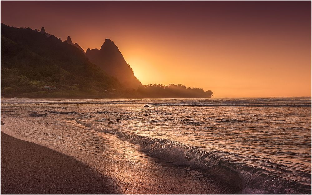 Peaceful coastal sunset illuminates beach and rocky cliffs in warm orange tones.
