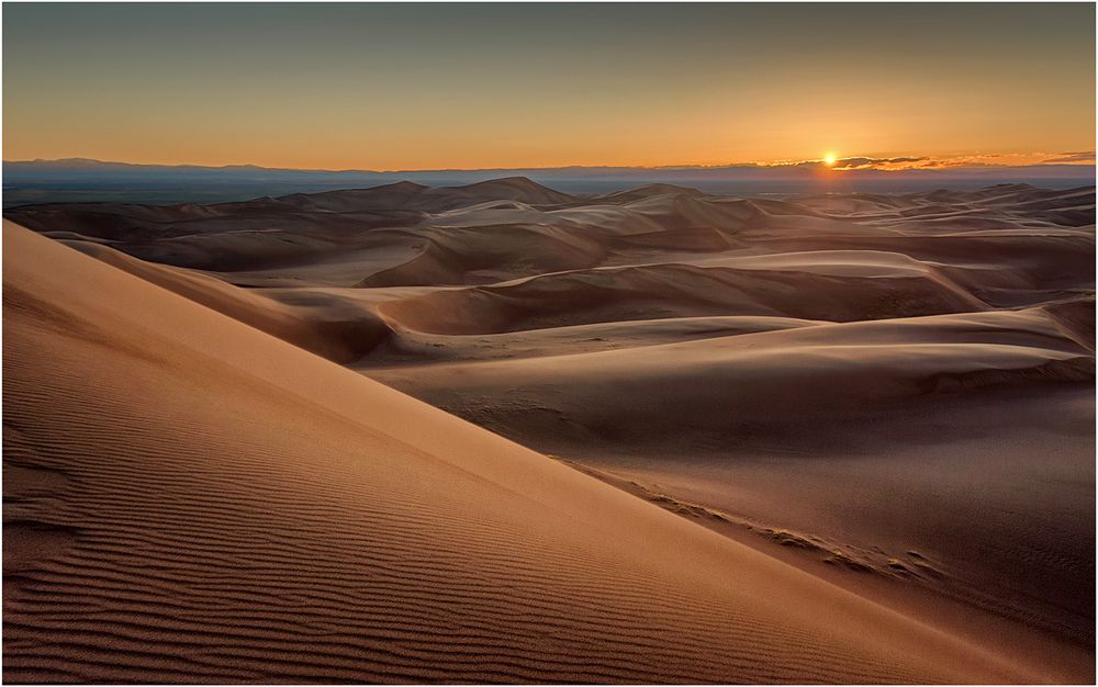 Rolling sand dunes stretch towards distant mountains under a warm desert sunset.