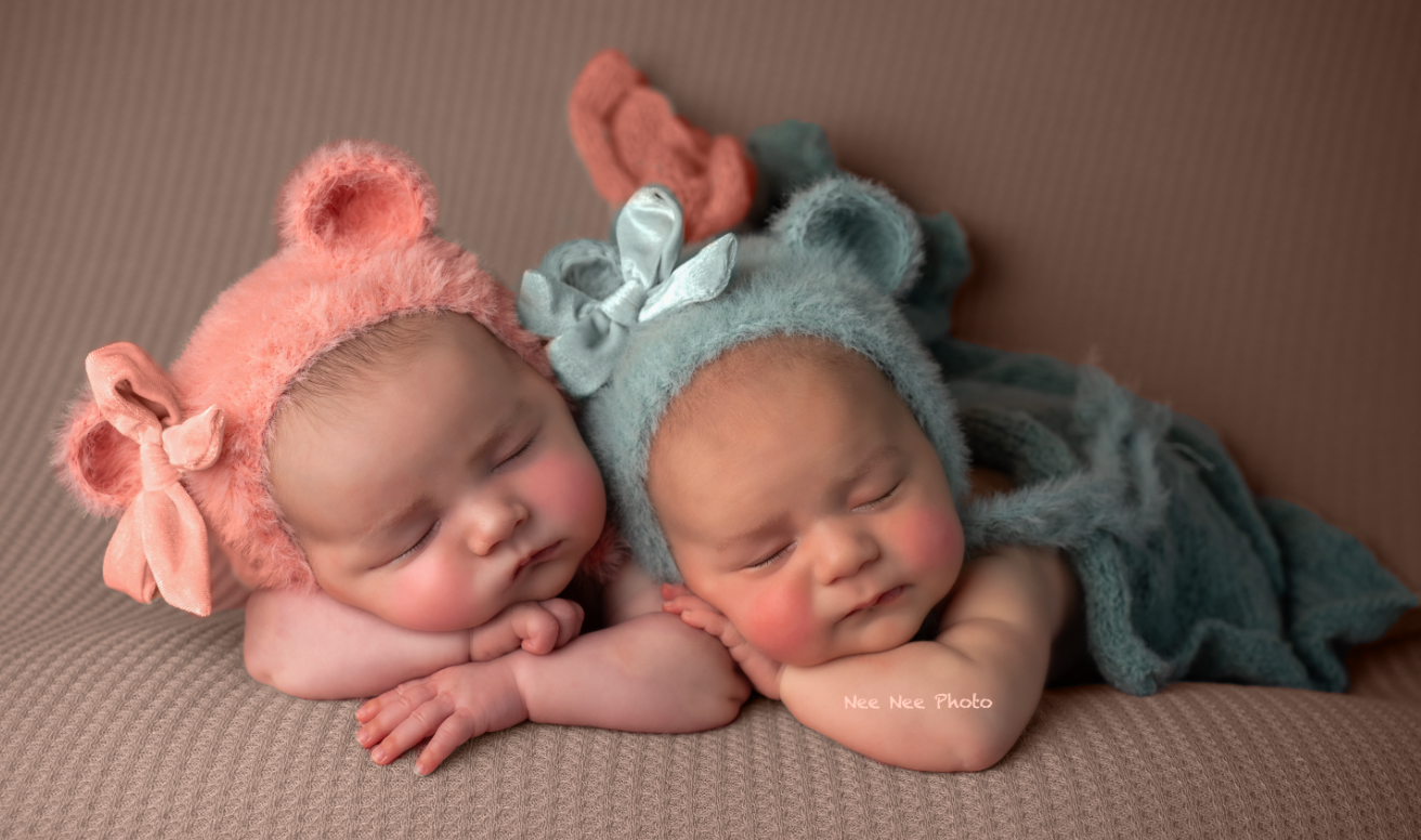 Two sleeping newborn twins wearing pink and blue bear bonnets posed together on a soft brown blanket.
