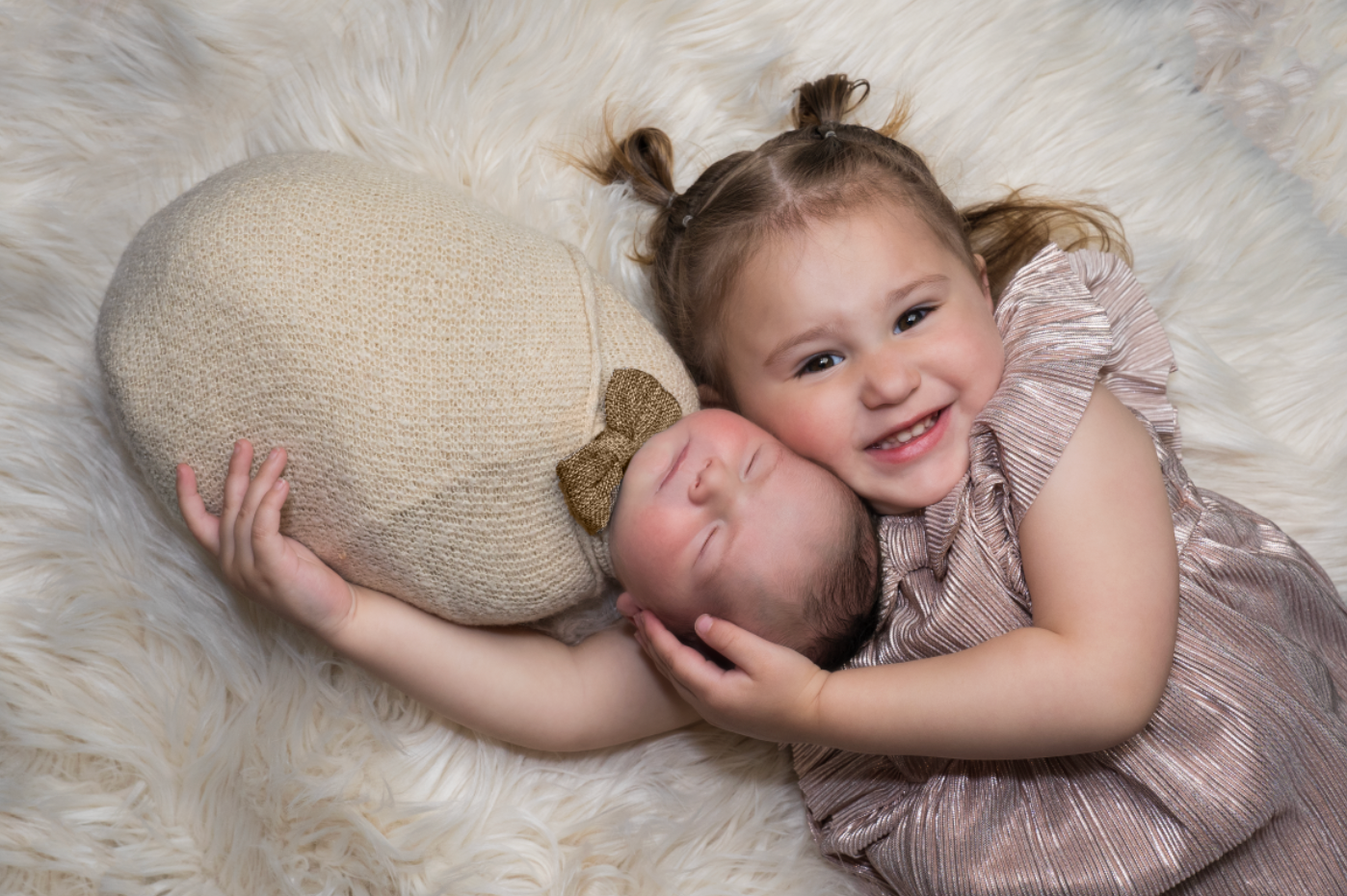 Smiling toddler girl in pink dress hugging sleeping newborn baby on fluffy white fur backdrop. Embracing Life’s Beauty
