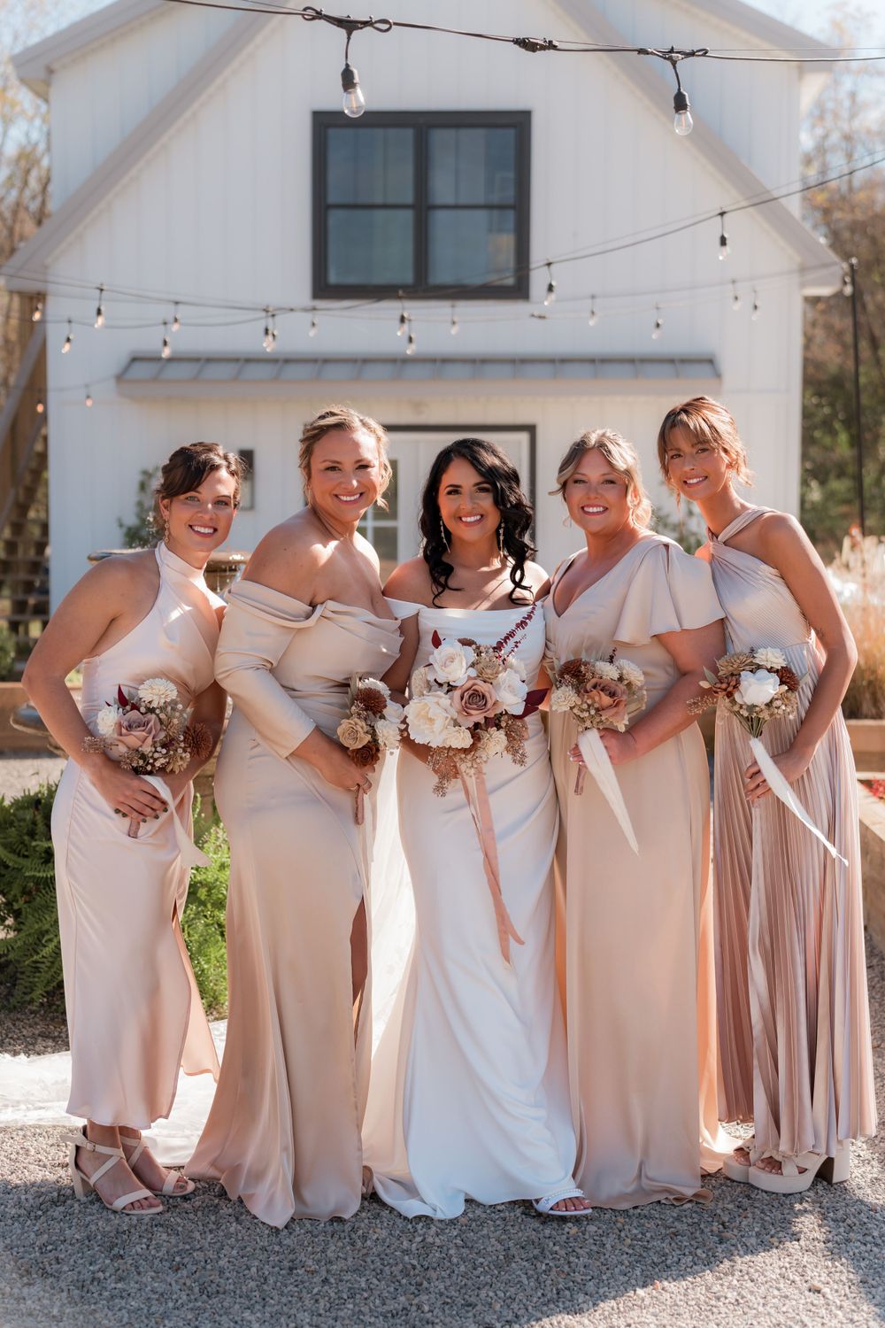 A bridal party in blush pink dresses poses together in front of a white building.