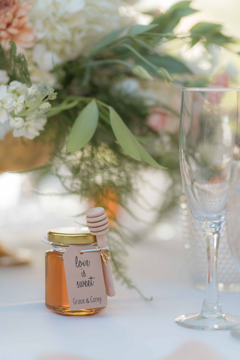 Small jar of honey wedding favor with floral centerpiece and champagne glass on white tablecloth.