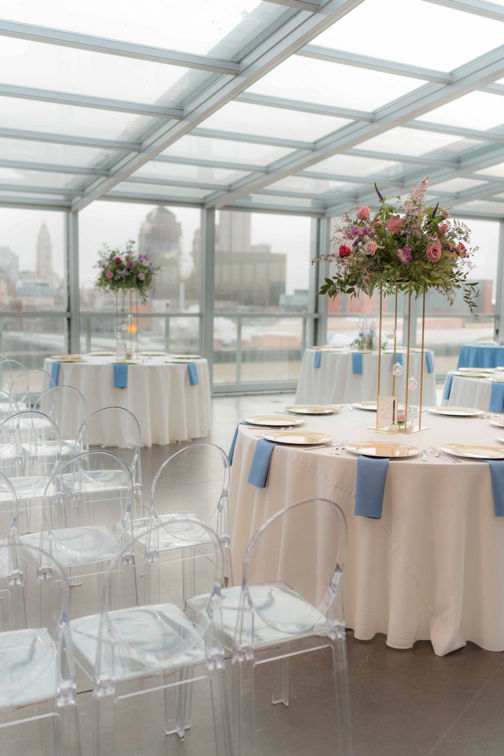 Modern reception venue with round tables, ghost chairs and tall floral centerpieces under glass ceiling.