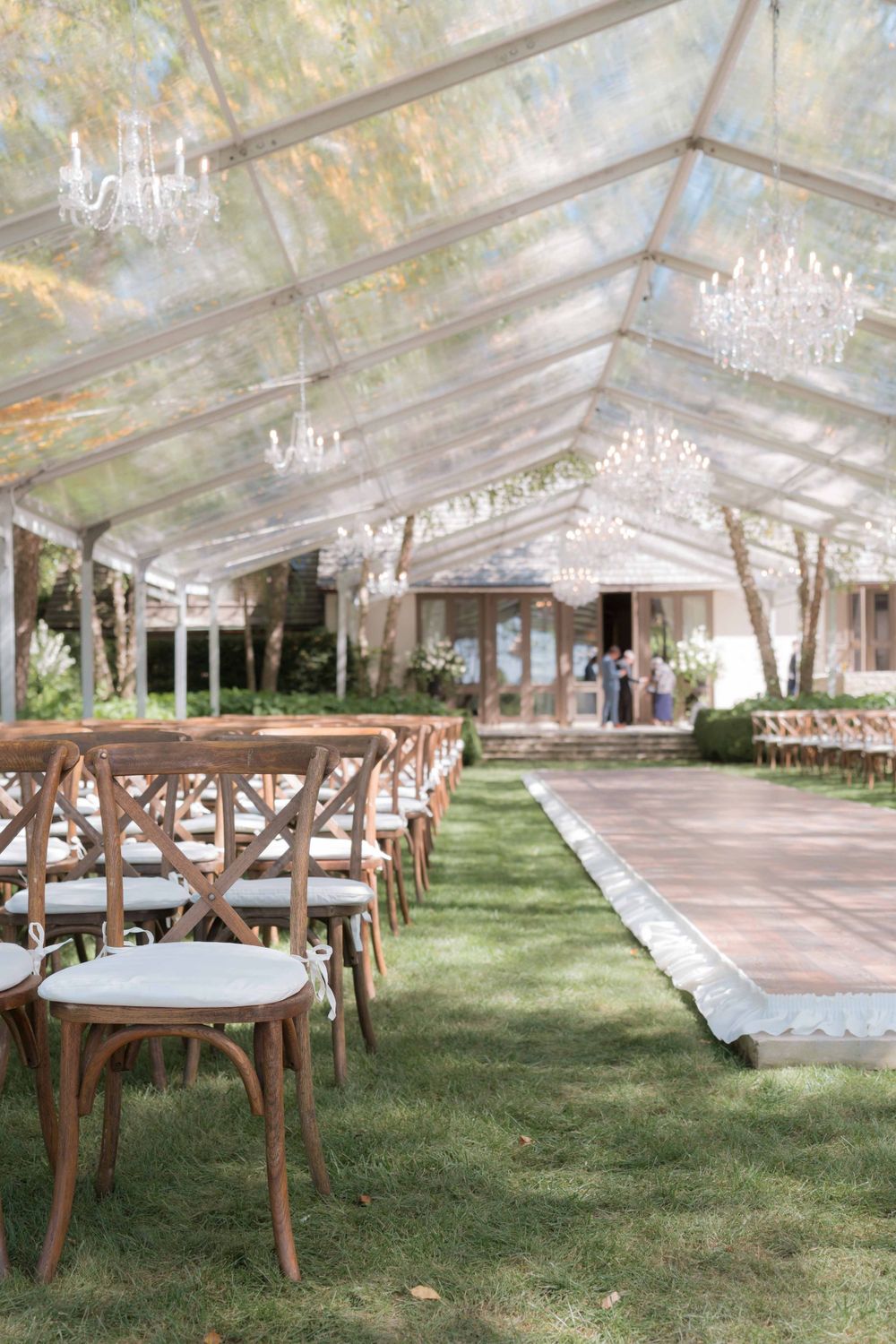 Elegant outdoor wedding ceremony setup with crystal chandeliers hanging from clear tent and wooden cross-back chairs.