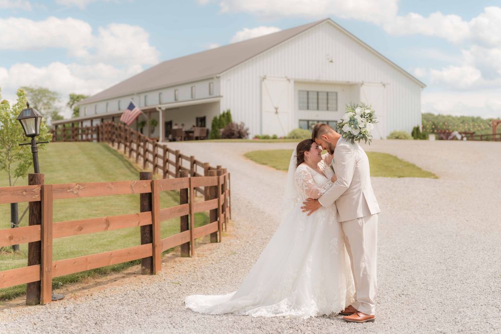 Bride and groom embrace outside white barn with wooden fence on sunny day.