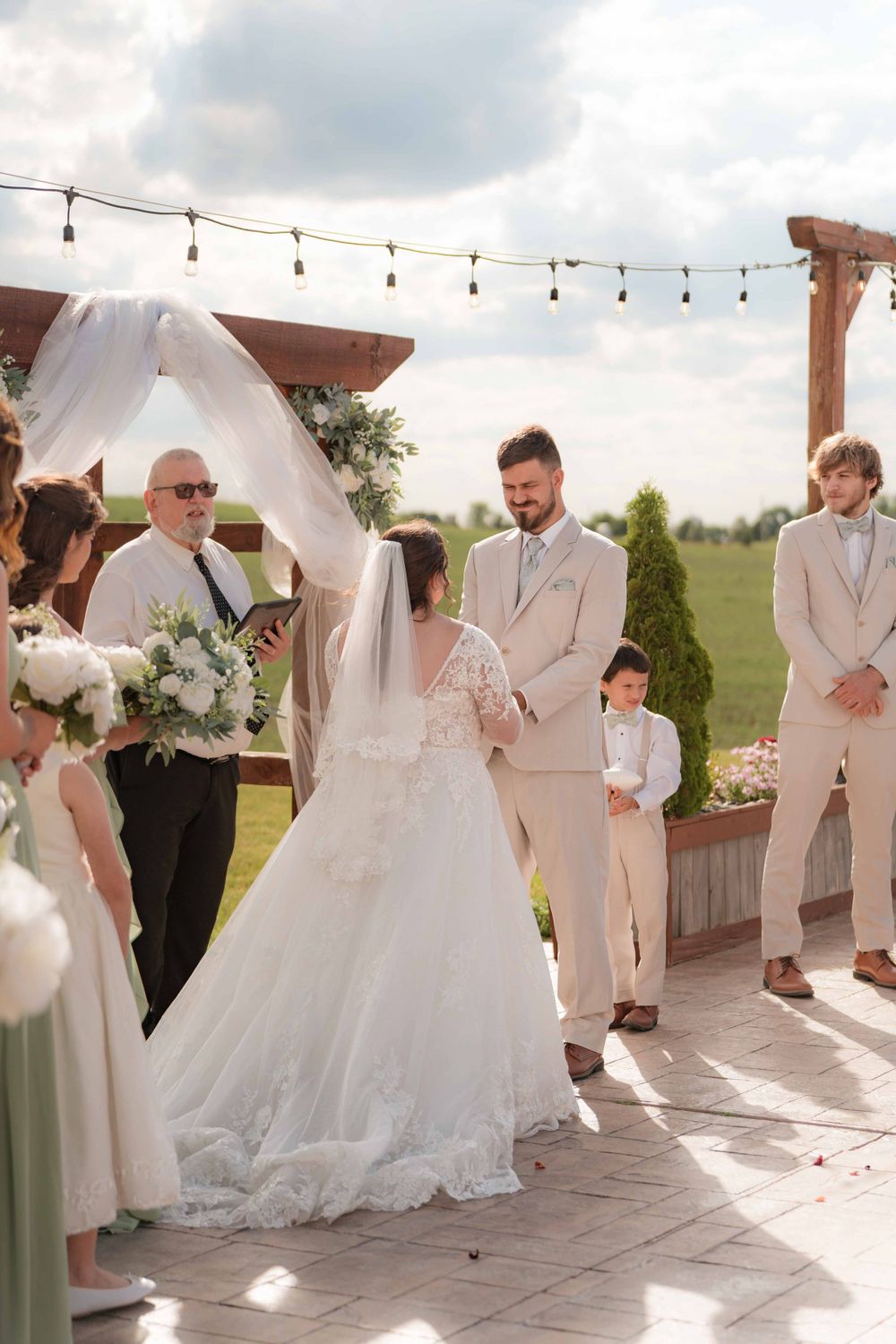 An outdoor wedding ceremony takes place under string lights with a white arch and floral decorations.