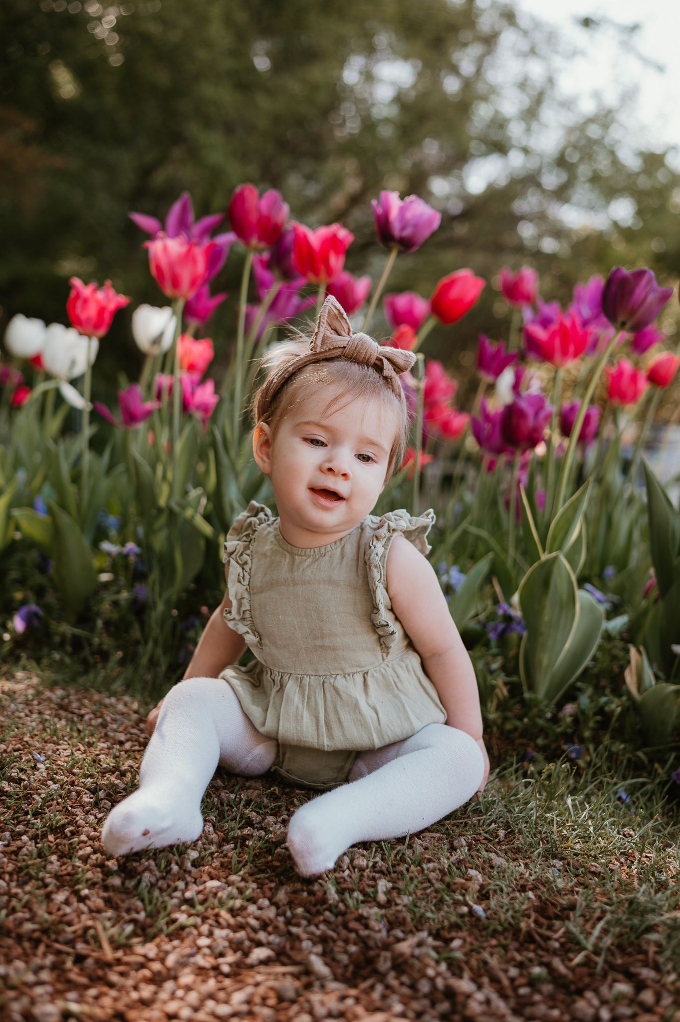 Baby in beige dress sitting among vibrant pink and purple tulips in a garden.