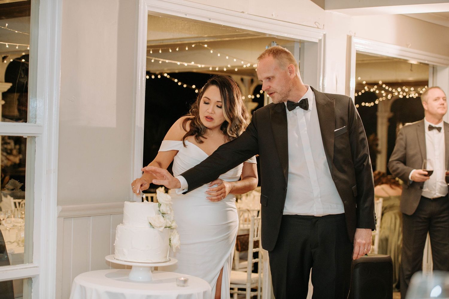 Couple cutting their elegant white wedding cake together at an indoor reception.
