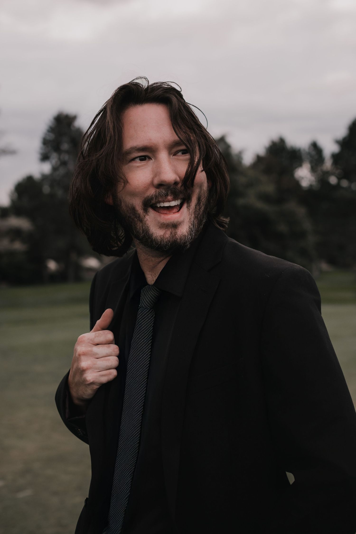 Joyful man with long dark hair wearing a black suit and tie laughs outdoors on an overcast day.