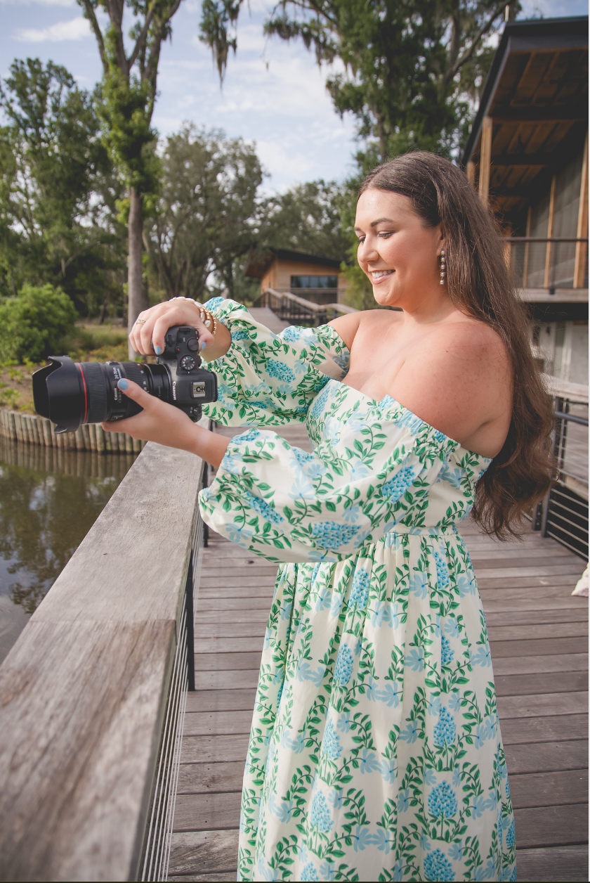 Person in floral dress holding camera on wooden deck overlooking water.