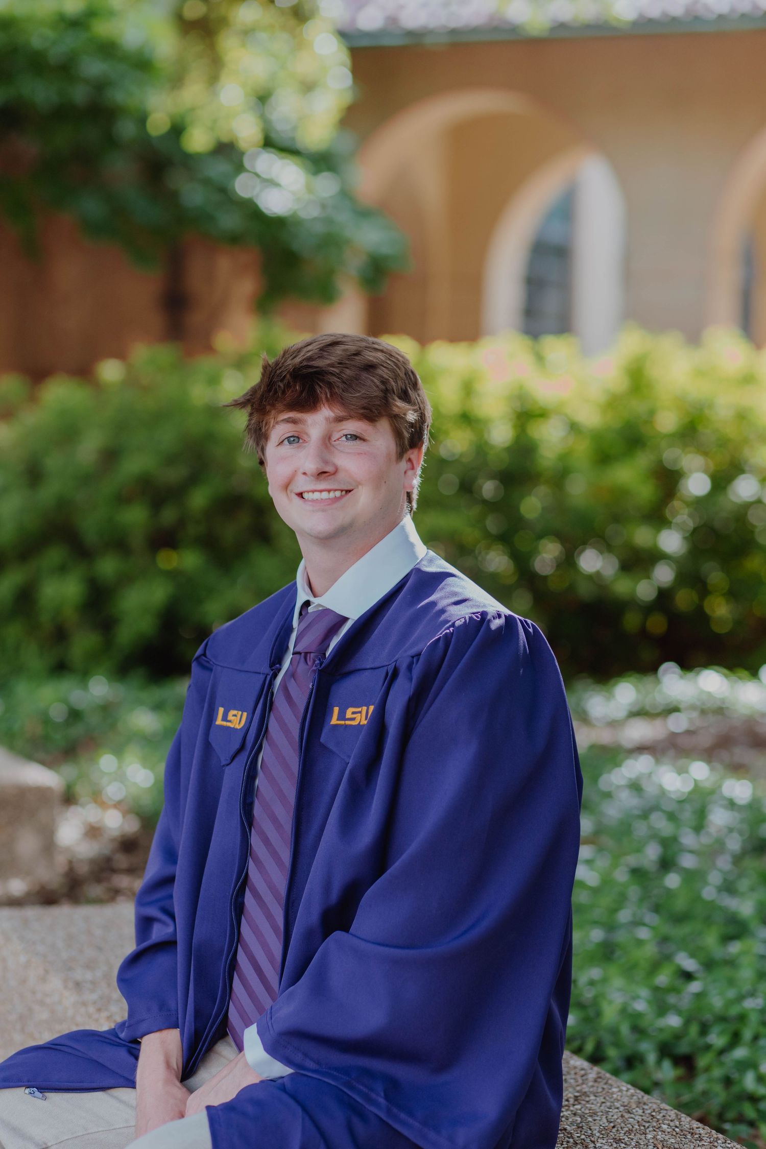 Graduate in blue cap and gown poses outside campus building in spring.