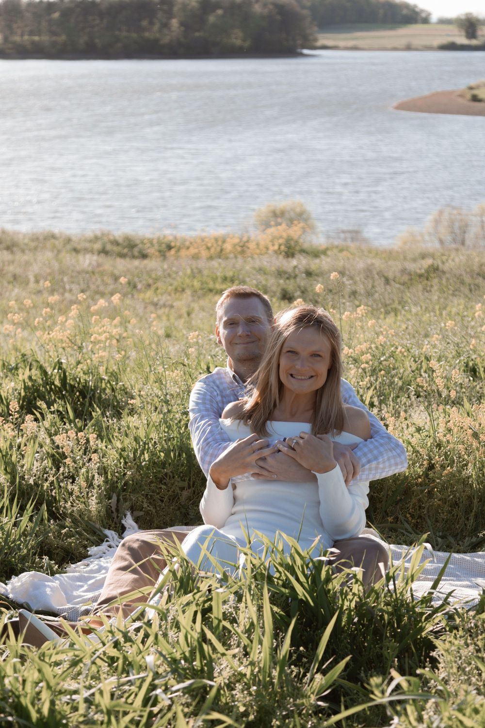 Couple relaxing on blanket by lakeside during sunny outdoor photoshoot.