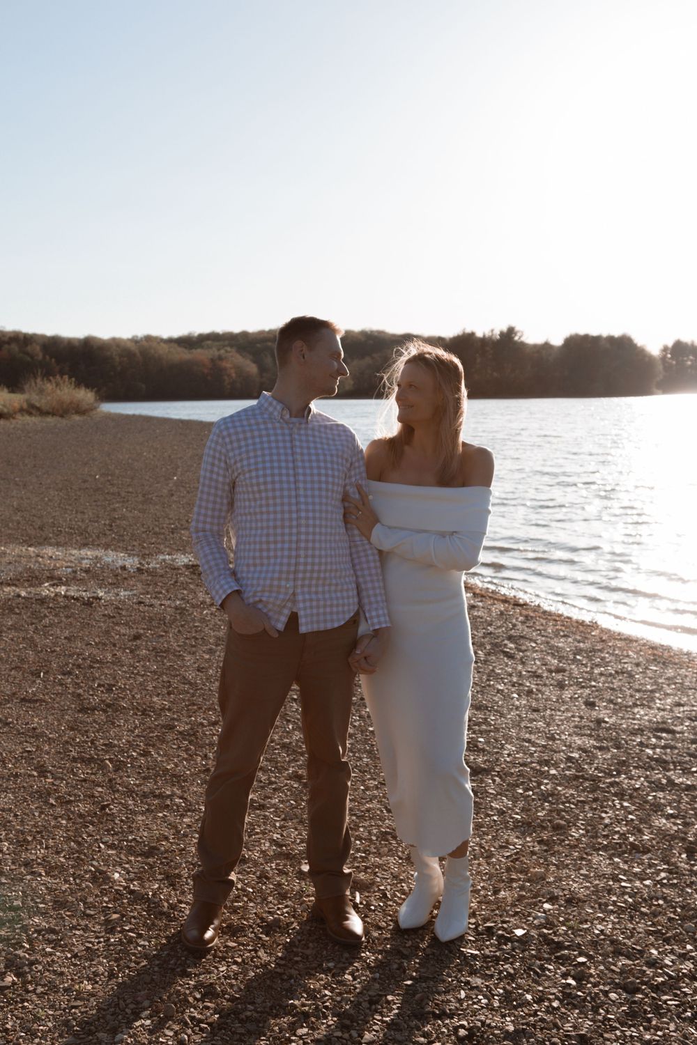 Couple stands together watching the sunset over a serene lake with trees in the background.