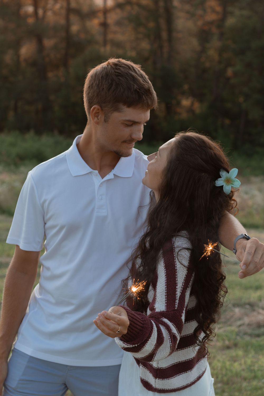 Romantic couple holding sparklers at sunset, wearing white polo and striped sweater with a flower in her hair.