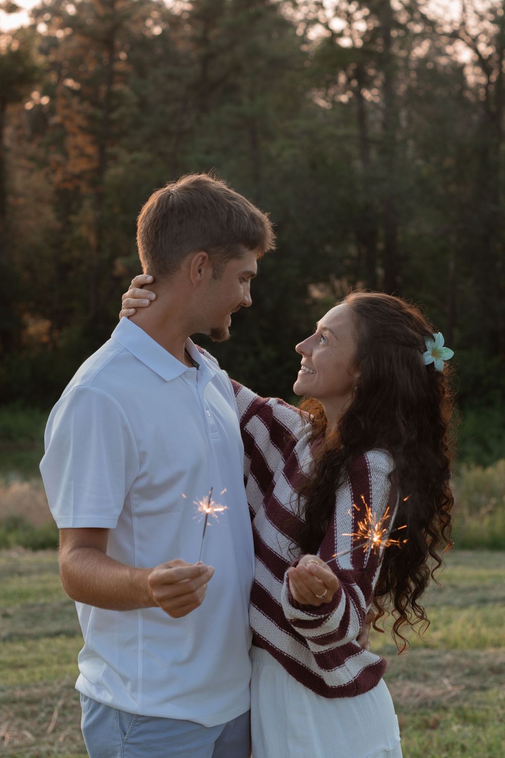 Romantic couple holding sparklers at sunset in a wooded field with warm golden lighting.