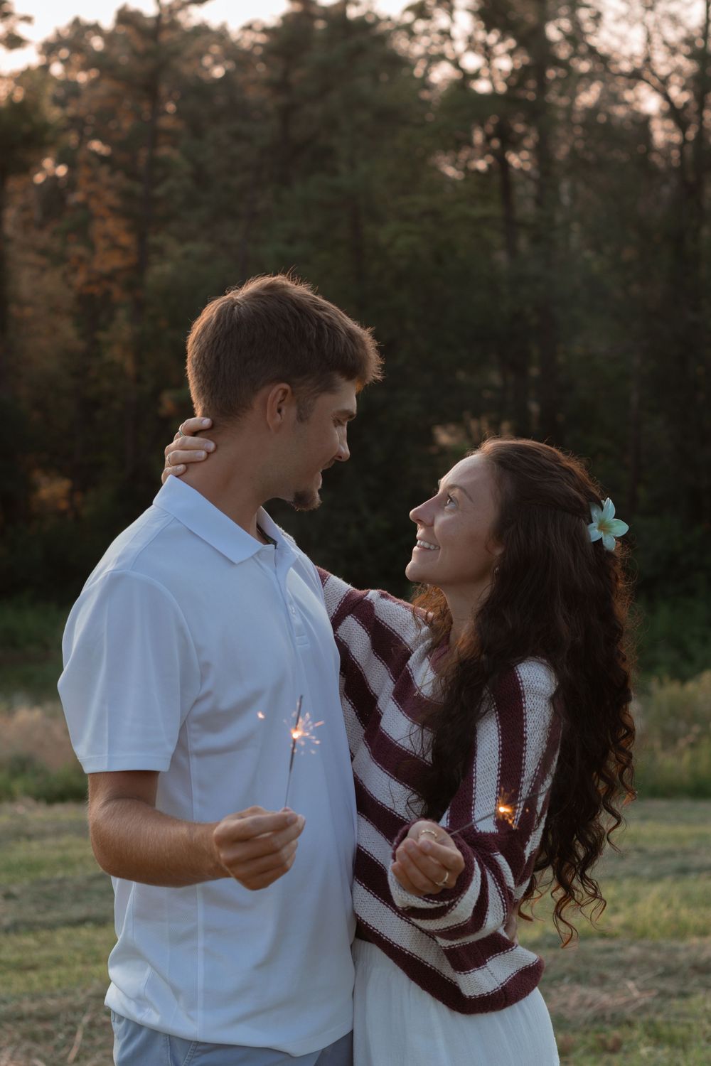 A romantic couple holds sparklers while sharing a joyful moment together at sunset in a wooded outdoor setting.