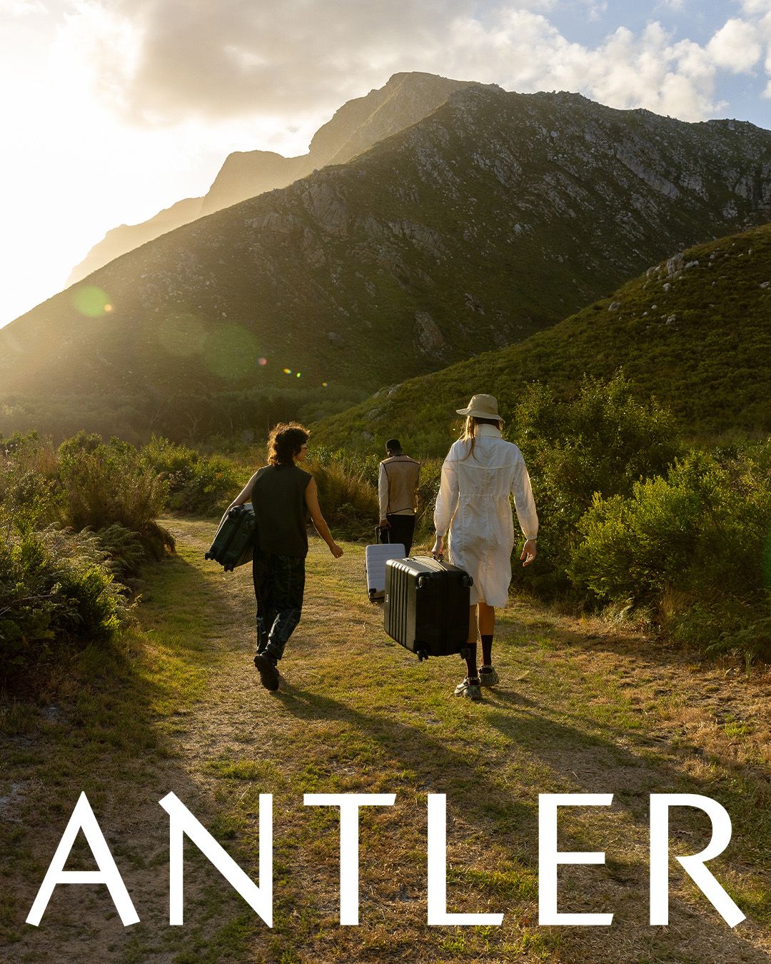 Three travelers walking along a mountain trail at sunset carrying luggage with Antler branding.
