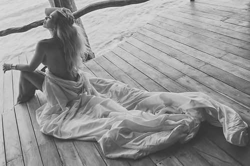 Black and white photo of bride in flowing dress sitting on wooden deck by ocean.