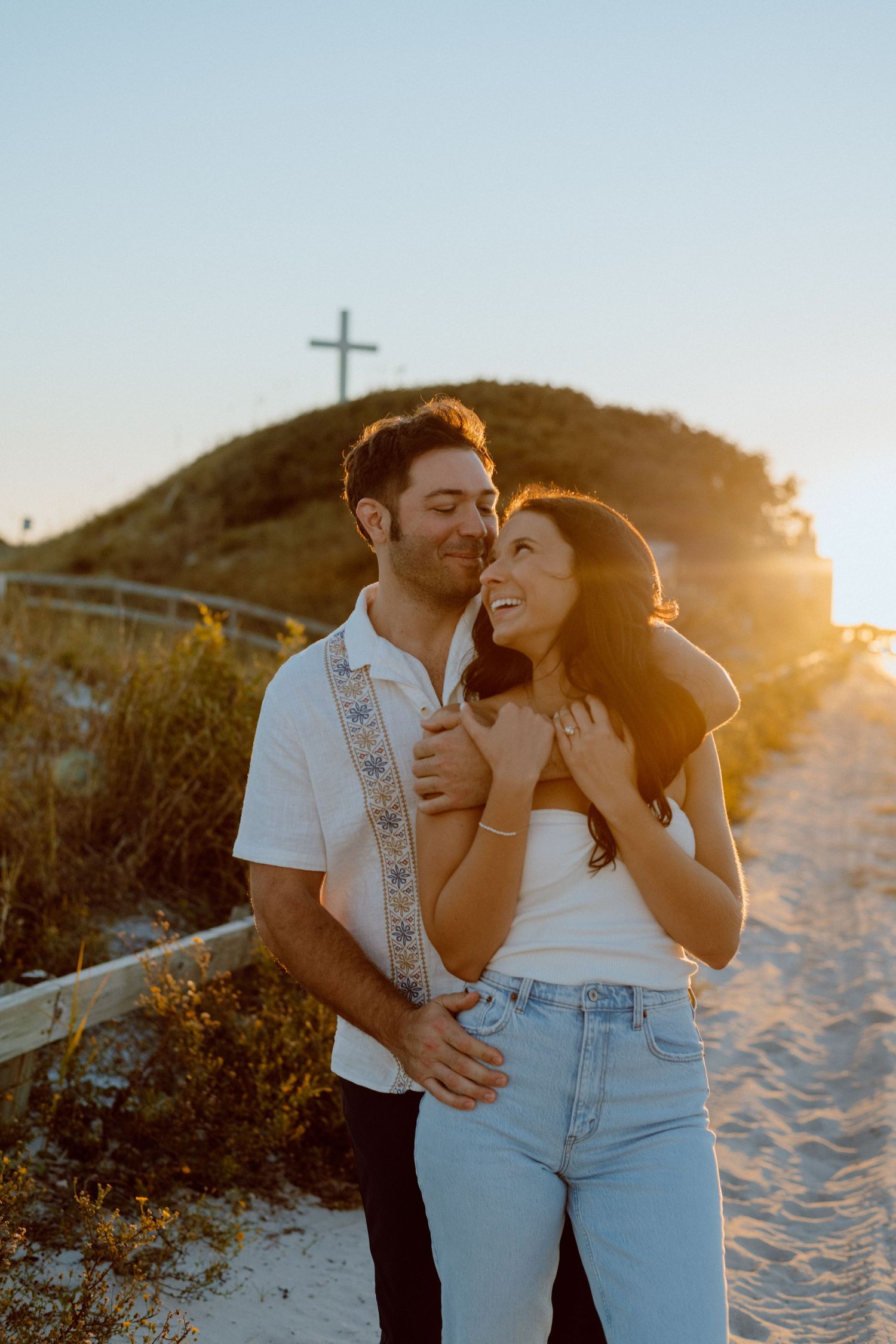 Romantic sunset engagement photo on sandy hill with cross silhouette in background.