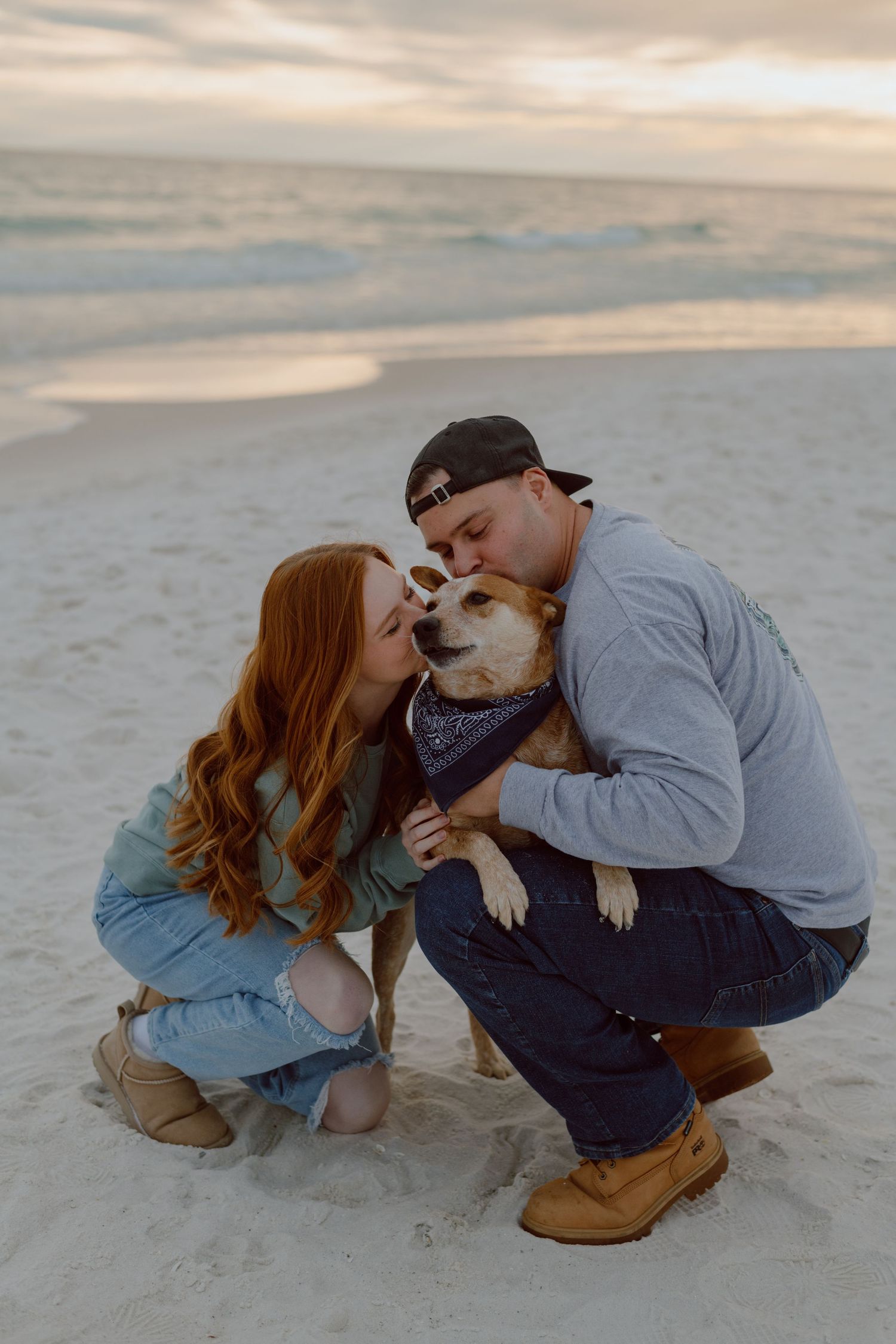Couple kneeling on beach sand to kiss their dog wearing a bandana at sunset.