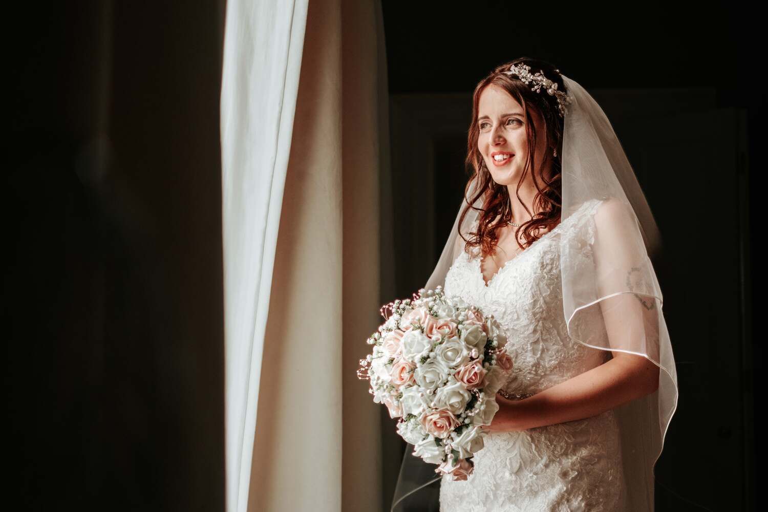 bride smiles and look out of window in full wedding gown holding flowers