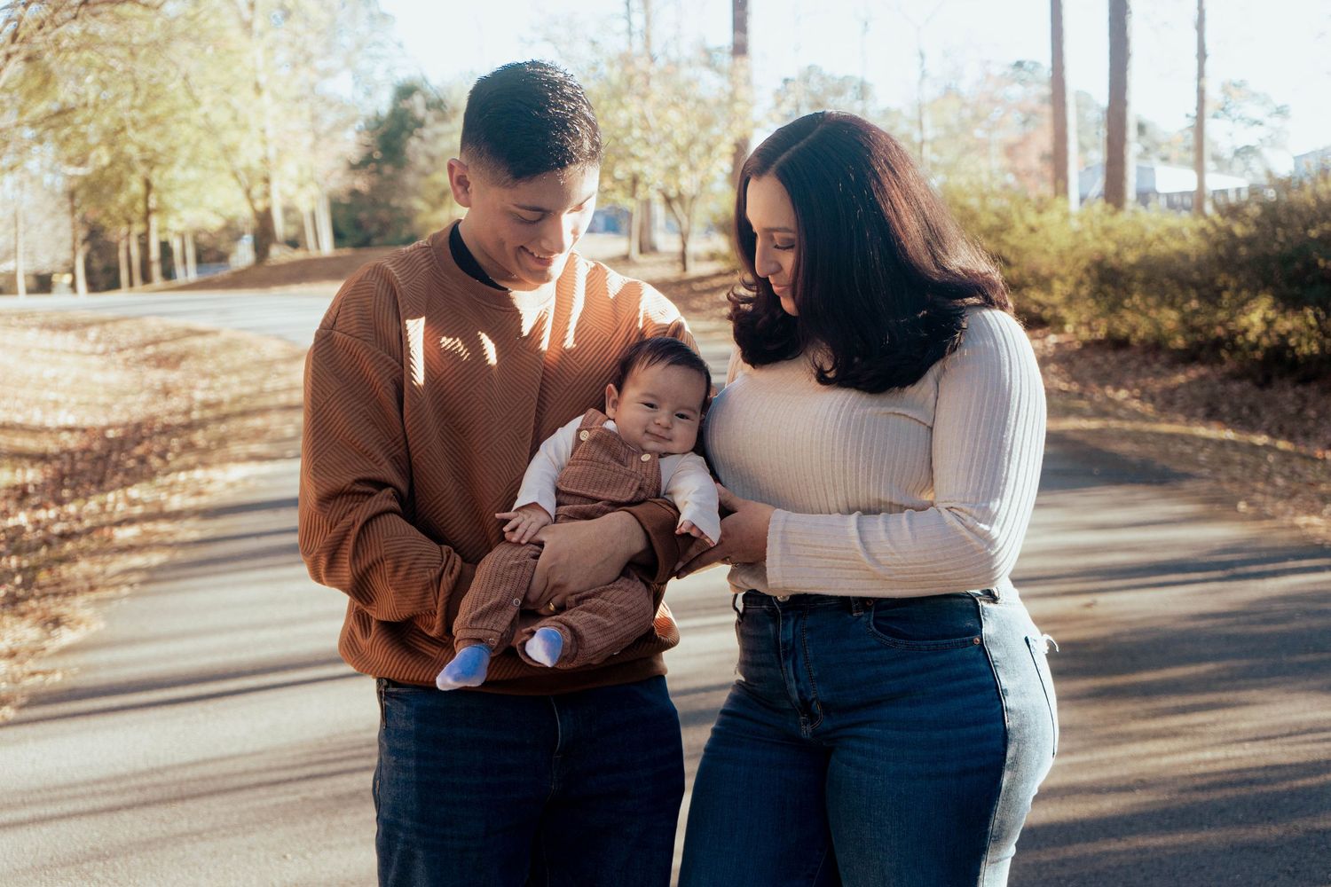 A couple stands together outdoors holding a small baby while walking along a path on an autumn day.