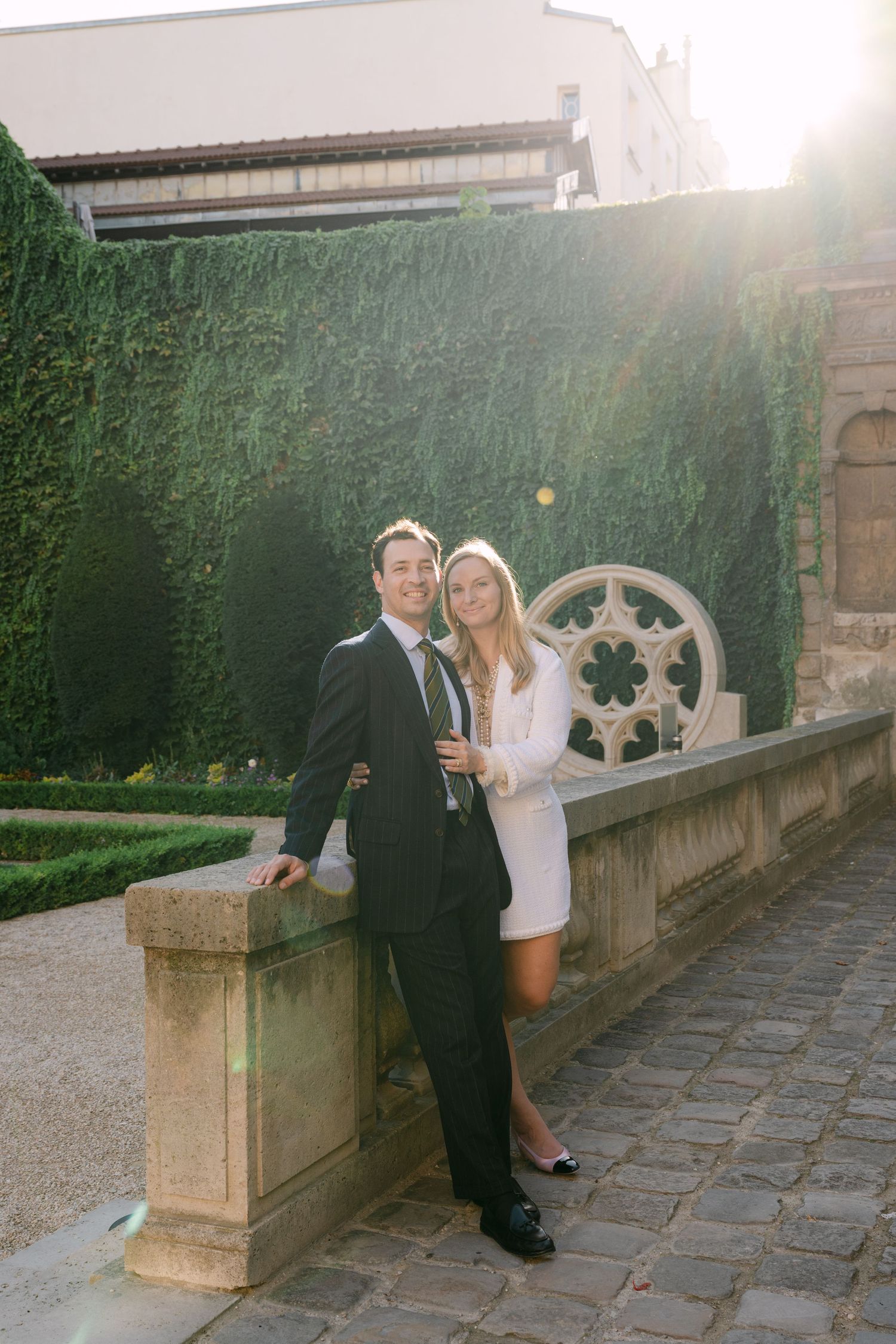A couple walks along a gravel path beside a classic French mansion with manicured hedges.