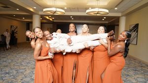 A group wearing peach-colored dresses poses together in a hotel hallway with elegant lighting and carpeting.