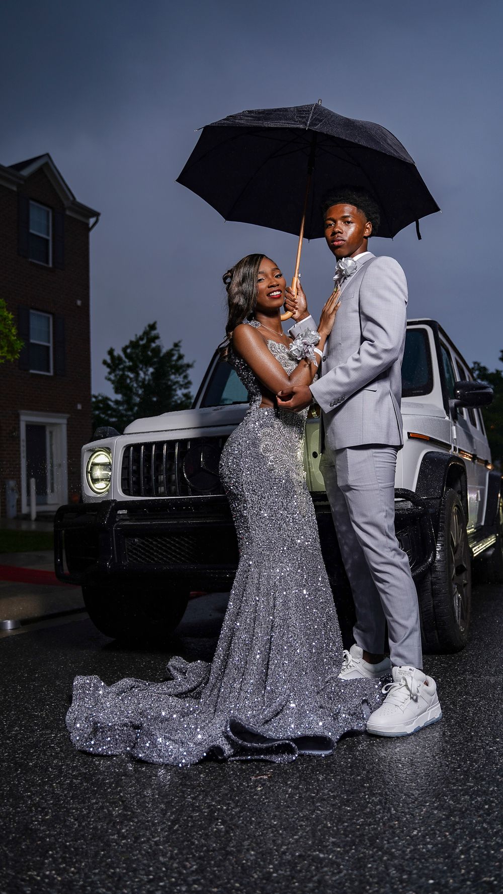 A couple shares a romantic moment under an umbrella in formal attire next to a vehicle on a rainy evening.