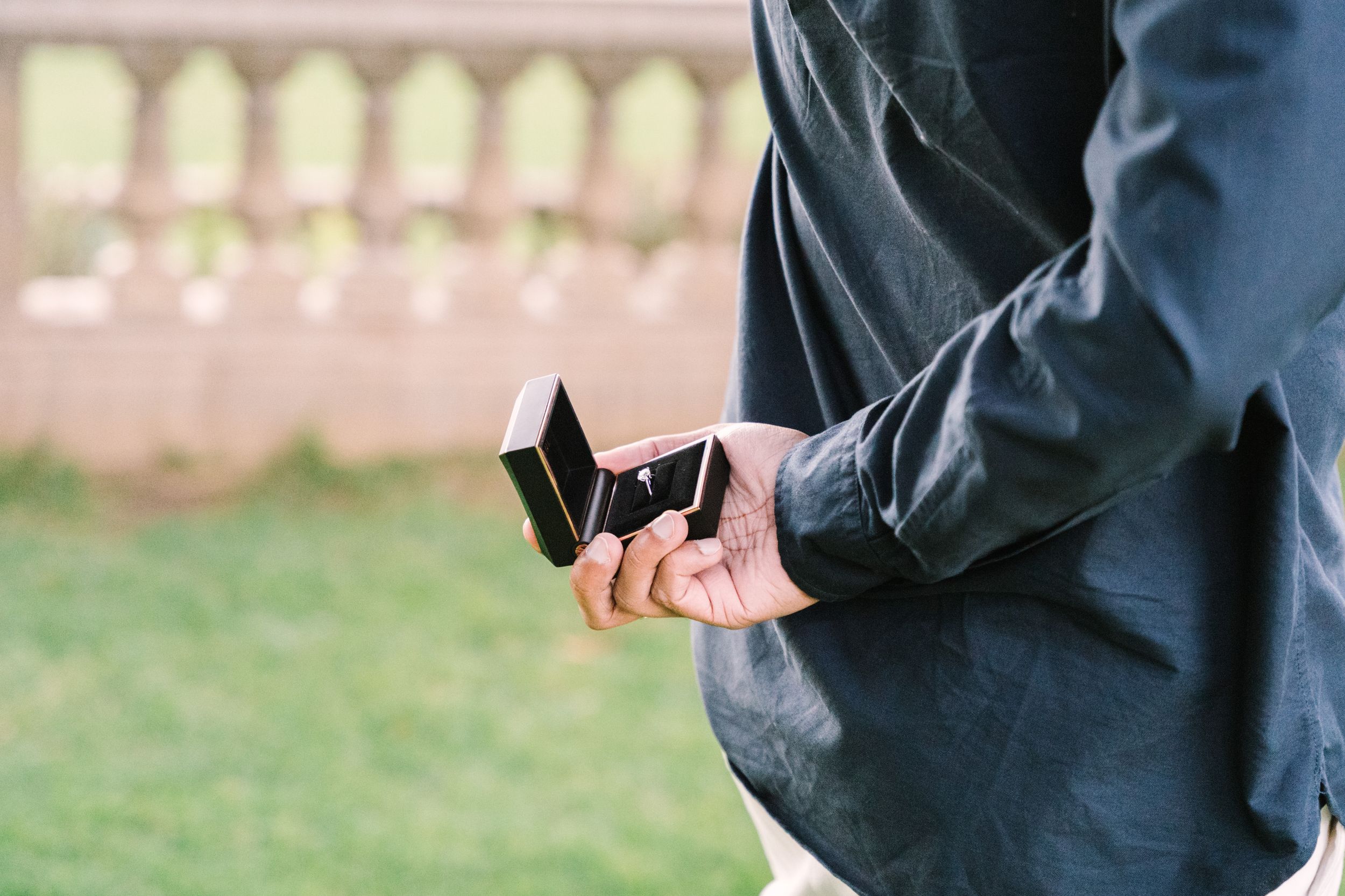 A man holding a ring in an open ring box behind his back wearing a blue shirt.