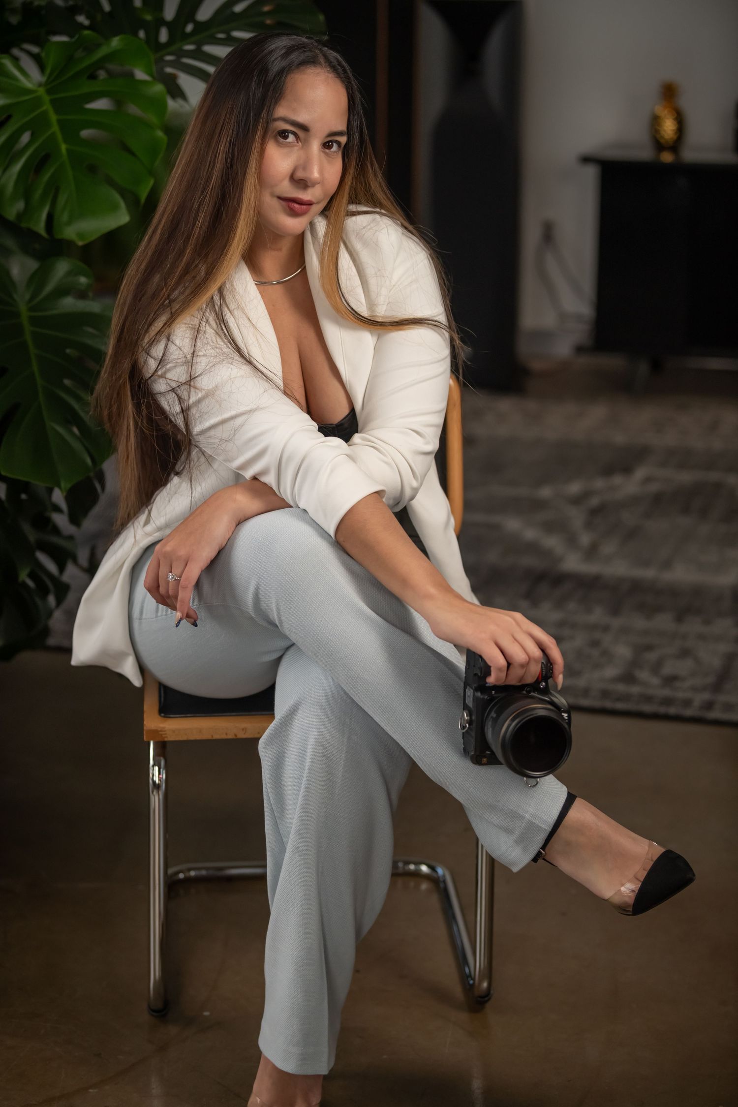 Elegant woman in white blazer and pants sitting on chair holding professional camera indoors.
