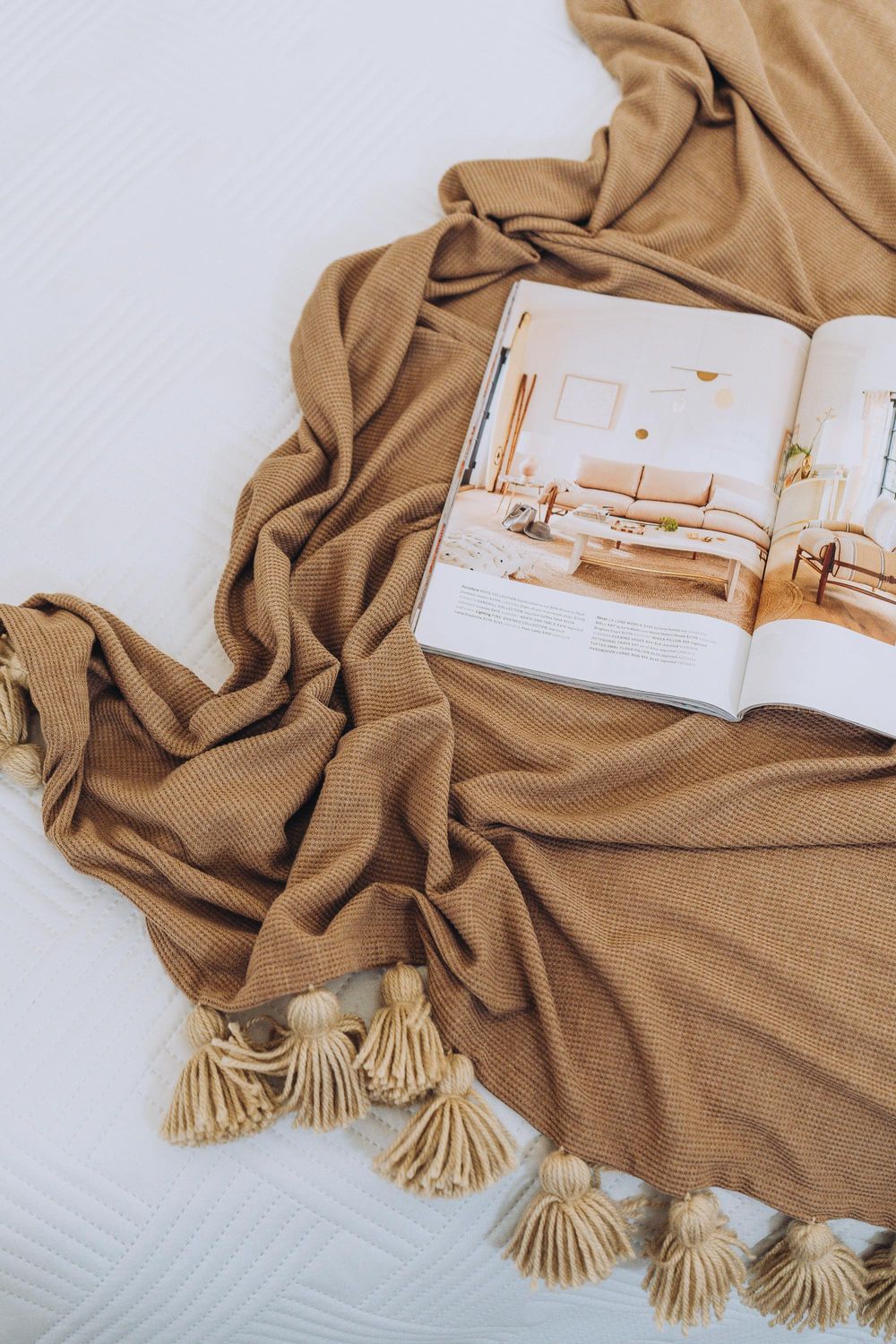 Tan throw blanket with tassels draped over open interior design magazine on white surface.