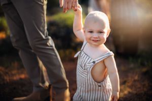 Toddler in striped outfit smiles while holding adult's hand in sunlit outdoor setting.
