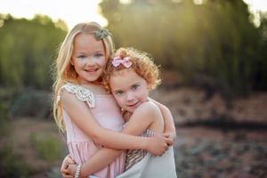 Two young children in light colored dresses hugging and smiling outdoors at sunset.