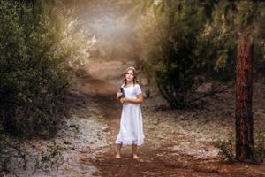 Child in white dress stands on dirt path in ethereal sunlit forest setting.