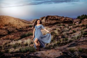 Woman in flowing blue dress twirling on desert rocks at sunset.