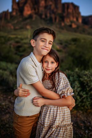 Two children embrace in front of red rock formations at golden hour in a desert landscape.