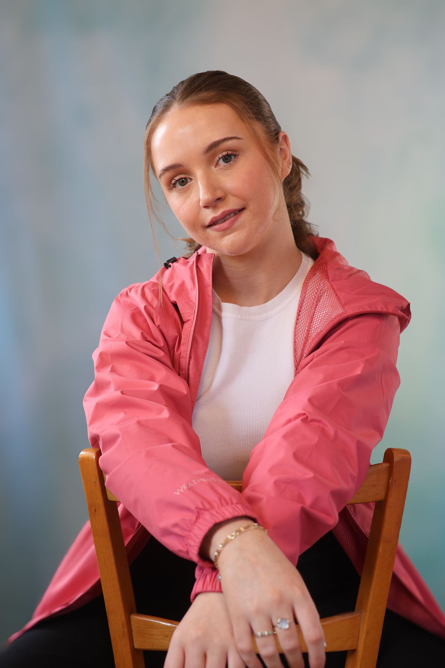 Person in pink windbreaker jacket sitting casually on wooden chair against blue background.