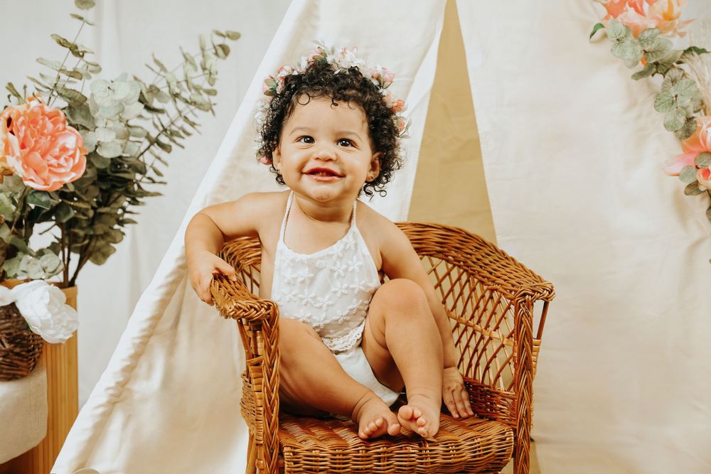 A toddler sits in a wicker chair with floral decor and white backdrop for a portrait session.