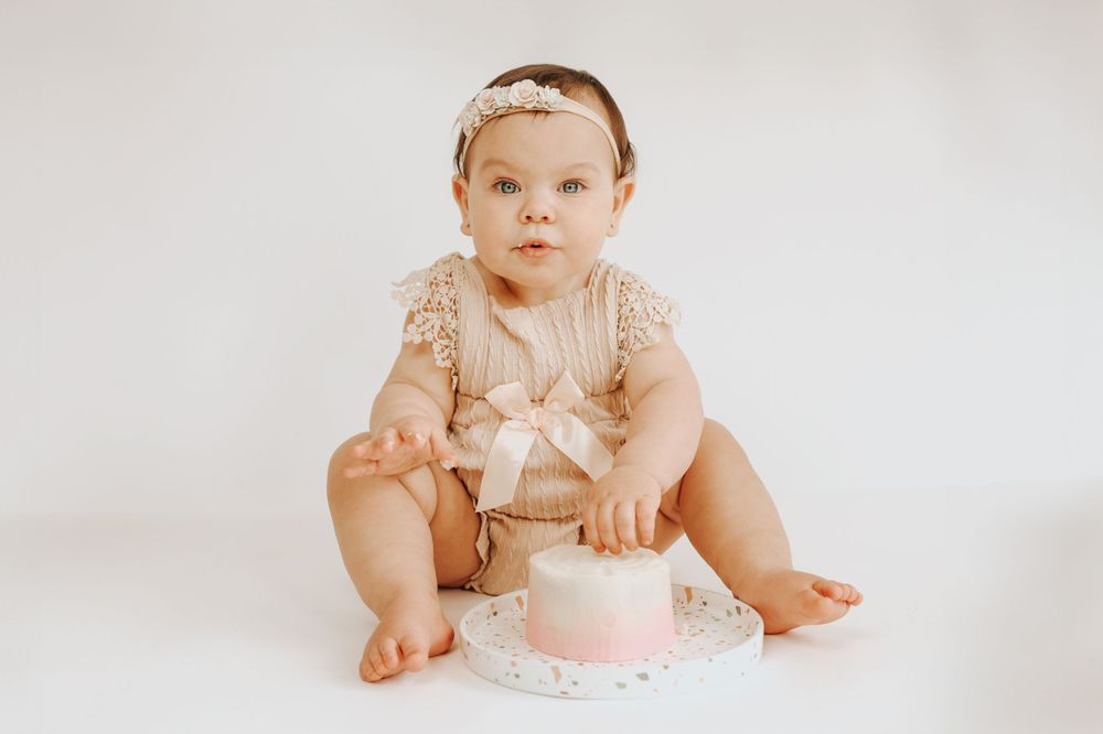 Infant wearing peach-colored lace outfit stretches on white backdrop during photo session.