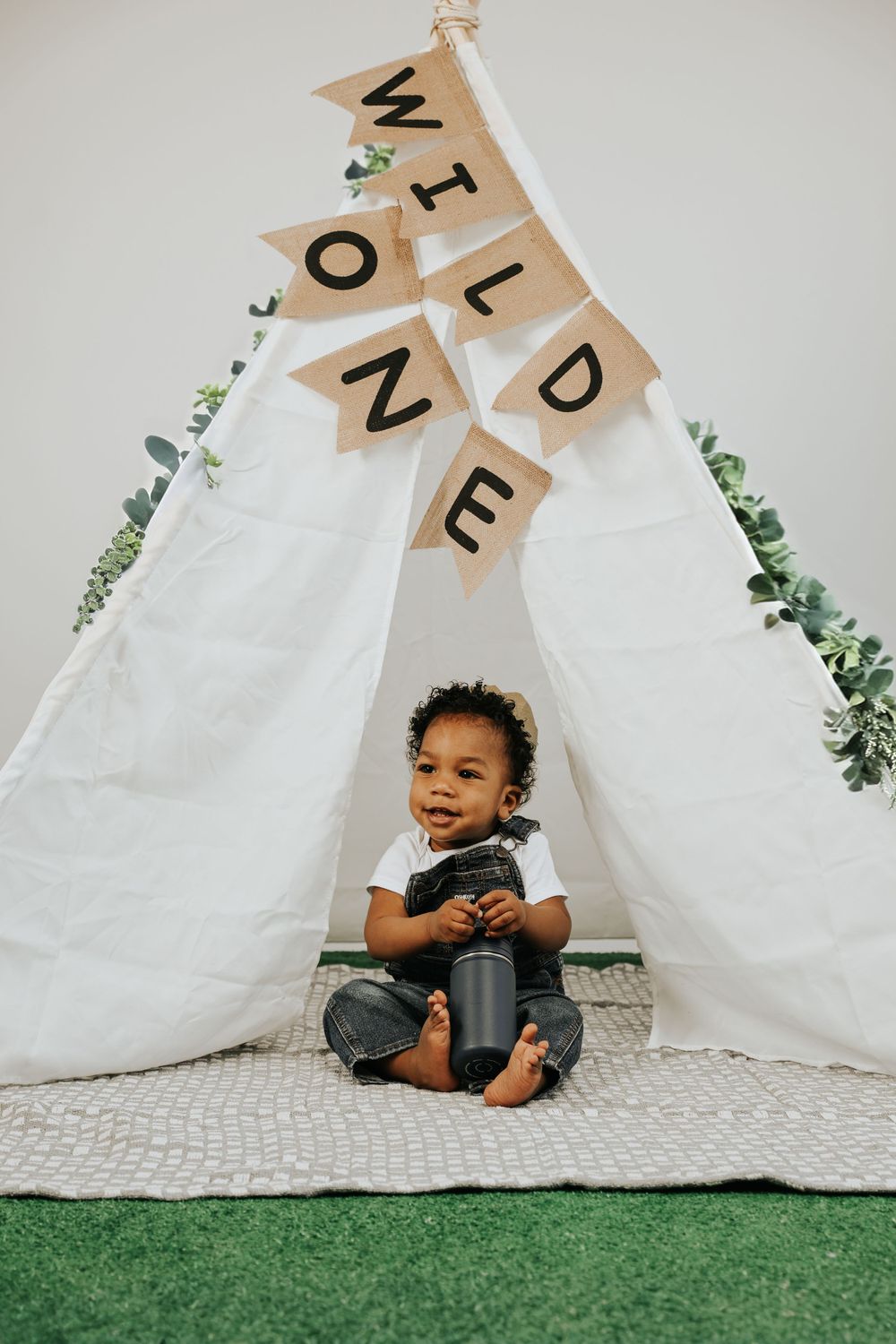 Child sits playfully in front of white teepee tent with decorative banner.