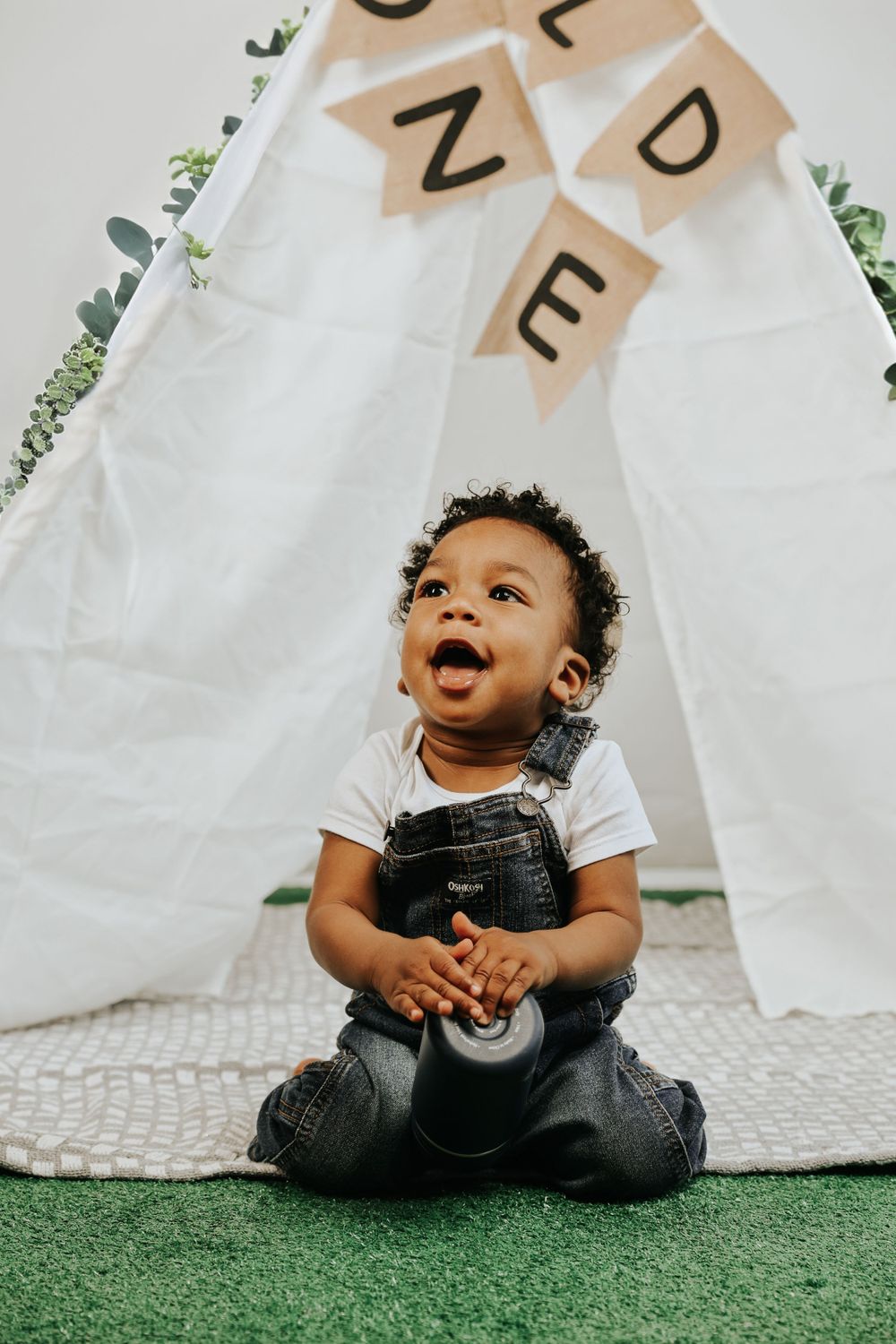 A toddler in denim overalls laughs joyfully while sitting on a carpet in front of a white tent.