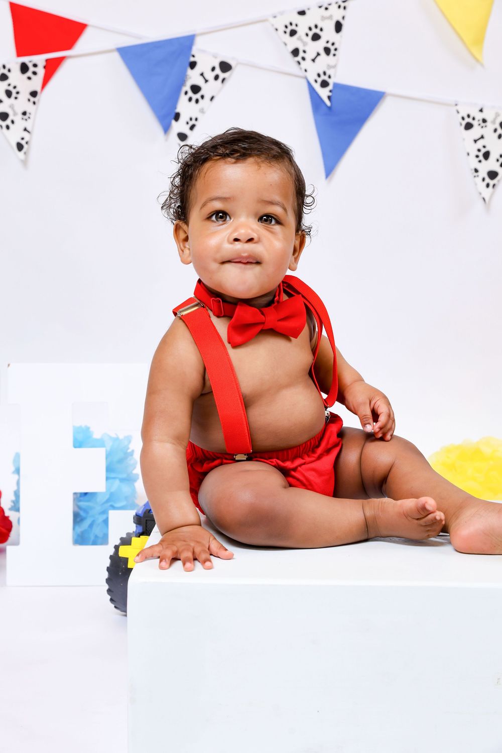 Baby in red bow tie outfit crawls on white floor under festive party banners.